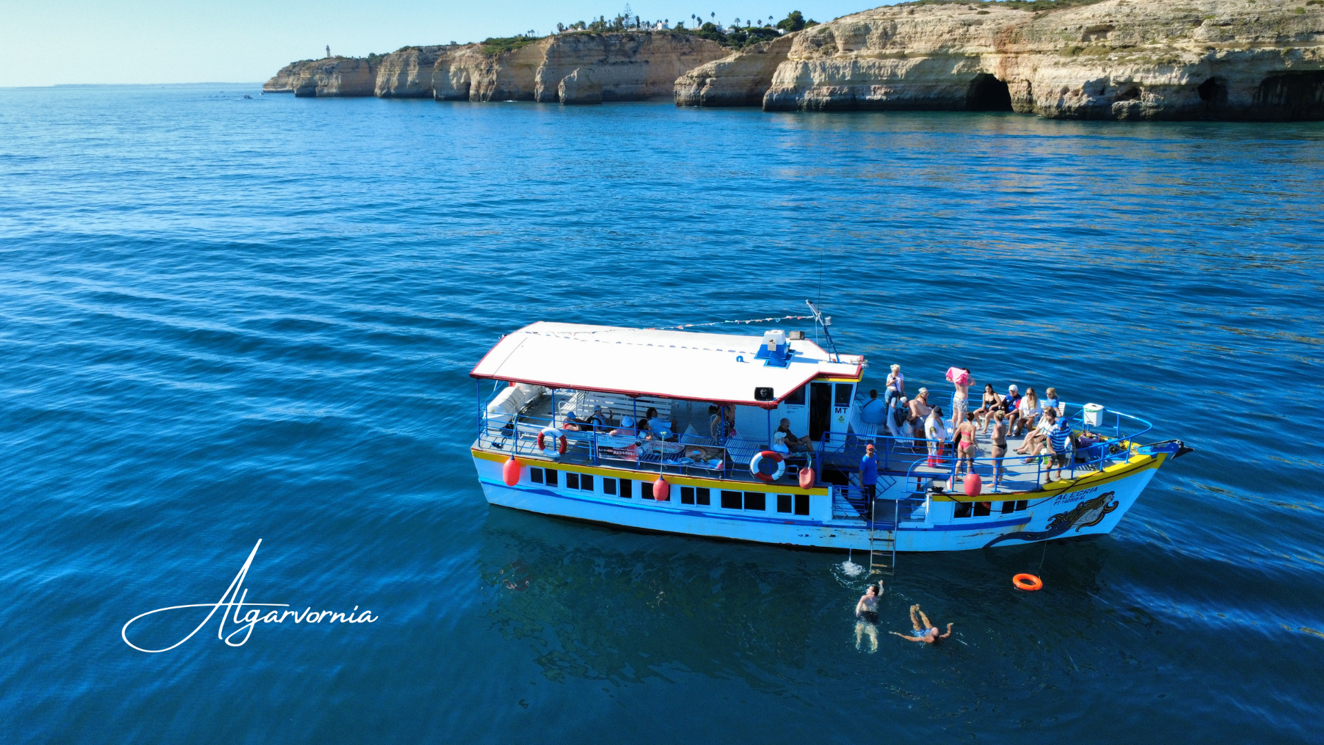 a tourism boat with people on boat and swimming in water with cliff coastline in background